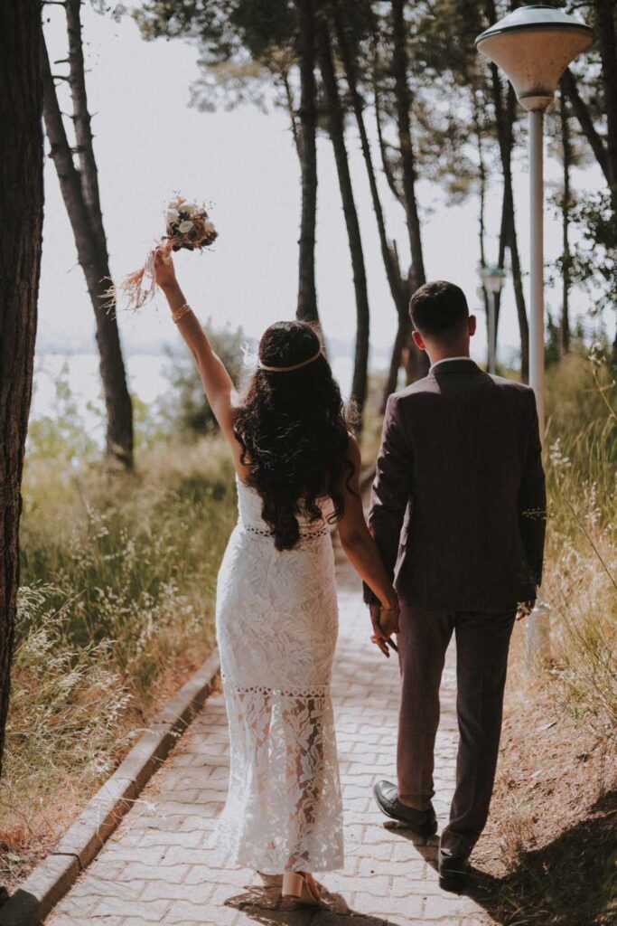 A couple, dressed formally for their couple photoshoot, walks hand in hand down a tree-lined path. The woman raises a bouquet into the air with her right hand.