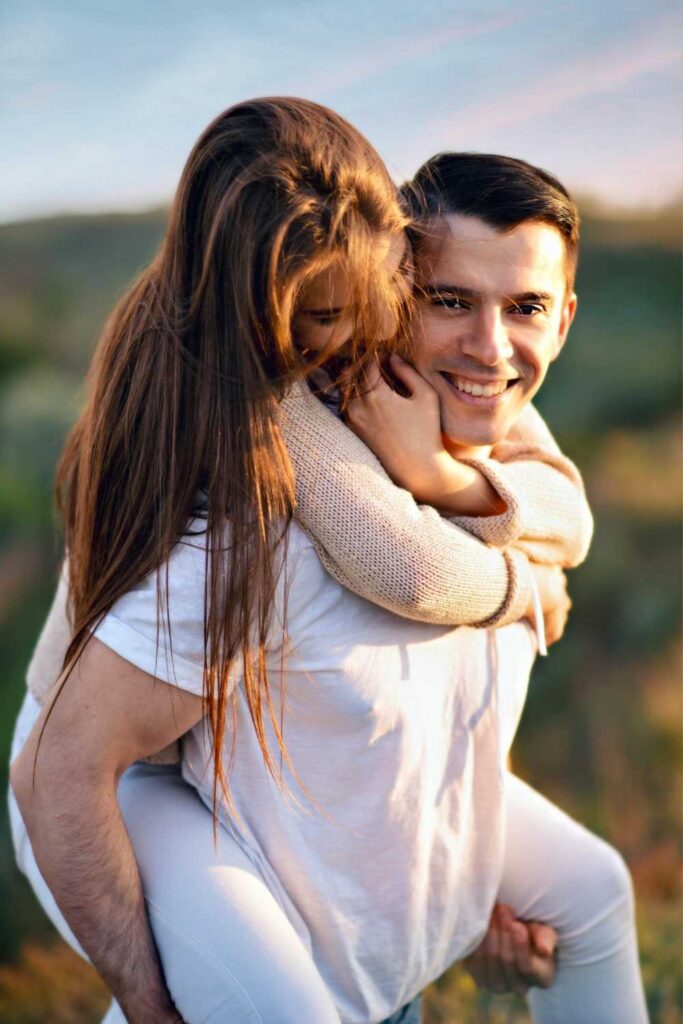 A man, wearing a white t-shirt, gives a piggyback ride to a woman with long brown hair in a white sweater and leggings. Both are smiling in an outdoor setting, capturing the joy of their couple photoshoot.