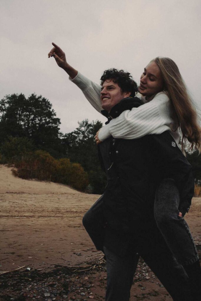 A man is giving a woman a piggyback ride on a beach. The woman is pointing forward, and they are both smiling. Trees and sand dunes are in the background on an overcast day, capturing a playful moment perfect for a couple photoshoot.