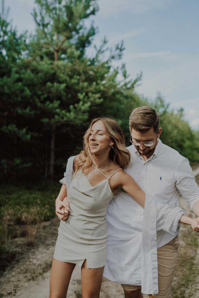 A couple in a beige dress and a white shirt walk joyfully along a dirt path, surrounded by greenery and trees, making for the perfect couple photoshoot.